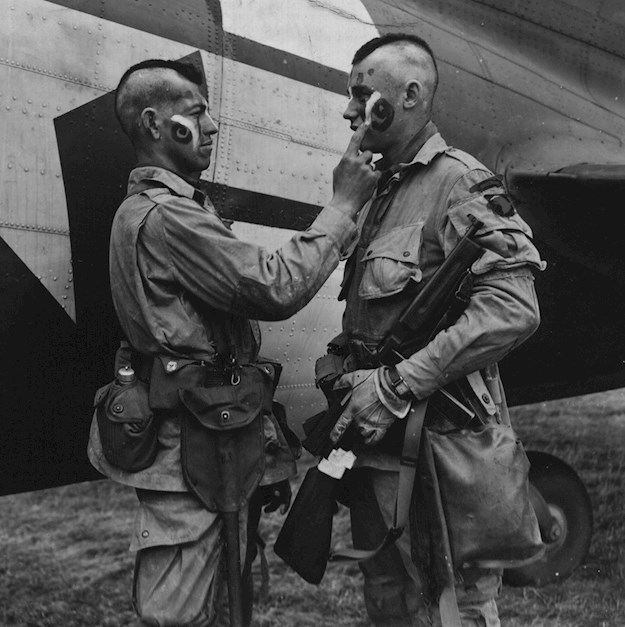 Private Clarence Ware applies war paint patterned after the American Indians to Private Charles Plaudo somewhere in England June 1944. © Creative Commons