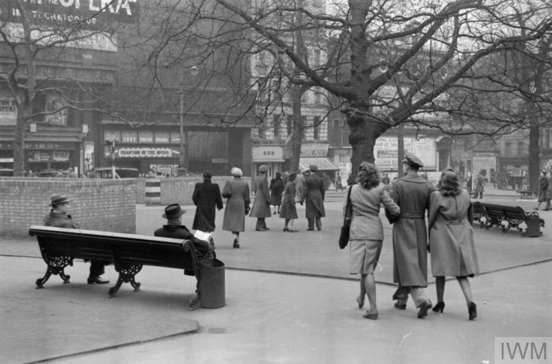 LONDON IN THE FIFTH YEAR OF WAR: EVERYDAY LIFE IN LONDON, ENGLAND, UK, 1944 (D 18309) A busy scene in London's Leicester Square.   An American soldier escorts two women past men resting on benches and a group of people chatting in the centre of the Square.  The trio are heading towards the Odeon Cinema, which is currently showing 'The Phantom of the Opera', starring Nelson Eddy, Susanna Foster and Claude Rains.  Also visible in the background are a newsagent's shop selling 'Picture... Copyright: © IWM. Original Source: http://www.iwm.org.uk/collections/item/object/205200673