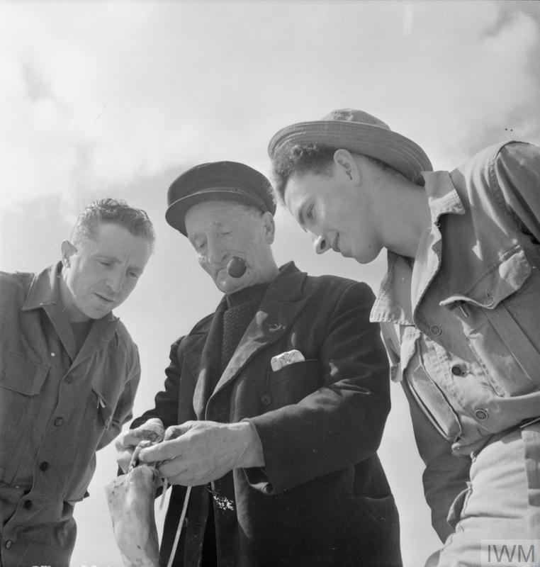 3- (c) IWUS TROOPS IN AN ENGLISH VILLAGE: EVERYDAY LIFE WITH THE AMERICANS IN BURTON BRADSTOCK, DORSET, ENGLAND, UK, 1944 (D 20132) Pfc Harris L Whitwell (left, of Main Street, Rogersville, Tennessee) and Pfc George W Burnett (of Route 5, Spartanburg, South Carolina) inspect a cuttle fish, held by local fisherman Tom Swaffield (centre).  The cuttlefish will be used in the lobster pots set by Tom, aided by the GIs.  Tom is smoking a pipe as he shows the soldiers the fish in the sunshine. Copyright: © IWM. Original Source: http://www.iwm.org.uk/collections/item/object/205200852M (D 20132)