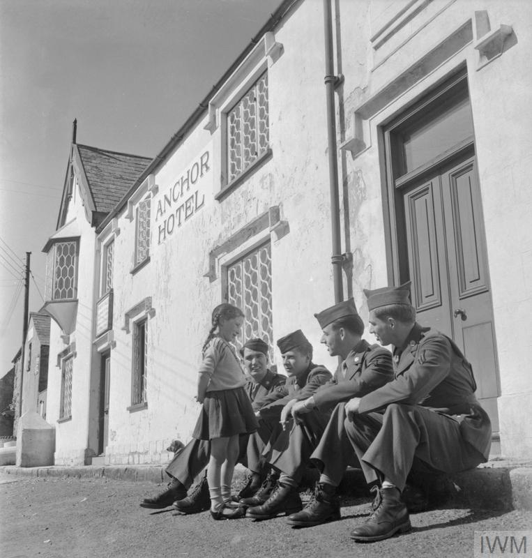 US TROOPS IN AN ENGLISH VILLAGE: EVERYDAY LIFE WITH THE AMERICANS IN BURTON BRADSTOCK, DORSET, ENGLAND, UK, 1944 (D 20133) Four American soldiers sit in the sunshine on the pavement outside The Anchor Inn in Burton Bradstock and chat to local girl Betty 'Freckles' Mackay.  According to the original caption, the nickname 'Freckles' was given to Betty by the GIs, who have 'made her a camp favourite'. Copyright: © IWM. Original Source: http://www.iwm.org.uk/collections/item/object/205200853