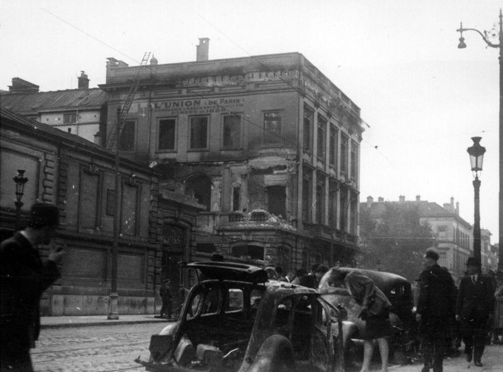 Burned out cars on rue de la Loi with, in the background, the fire-damaged buildings of the insurance company L'Union